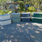 Four empty cascading metal raised garden beds, each showing the color options sitting on gravel in front of a garden landscape. 