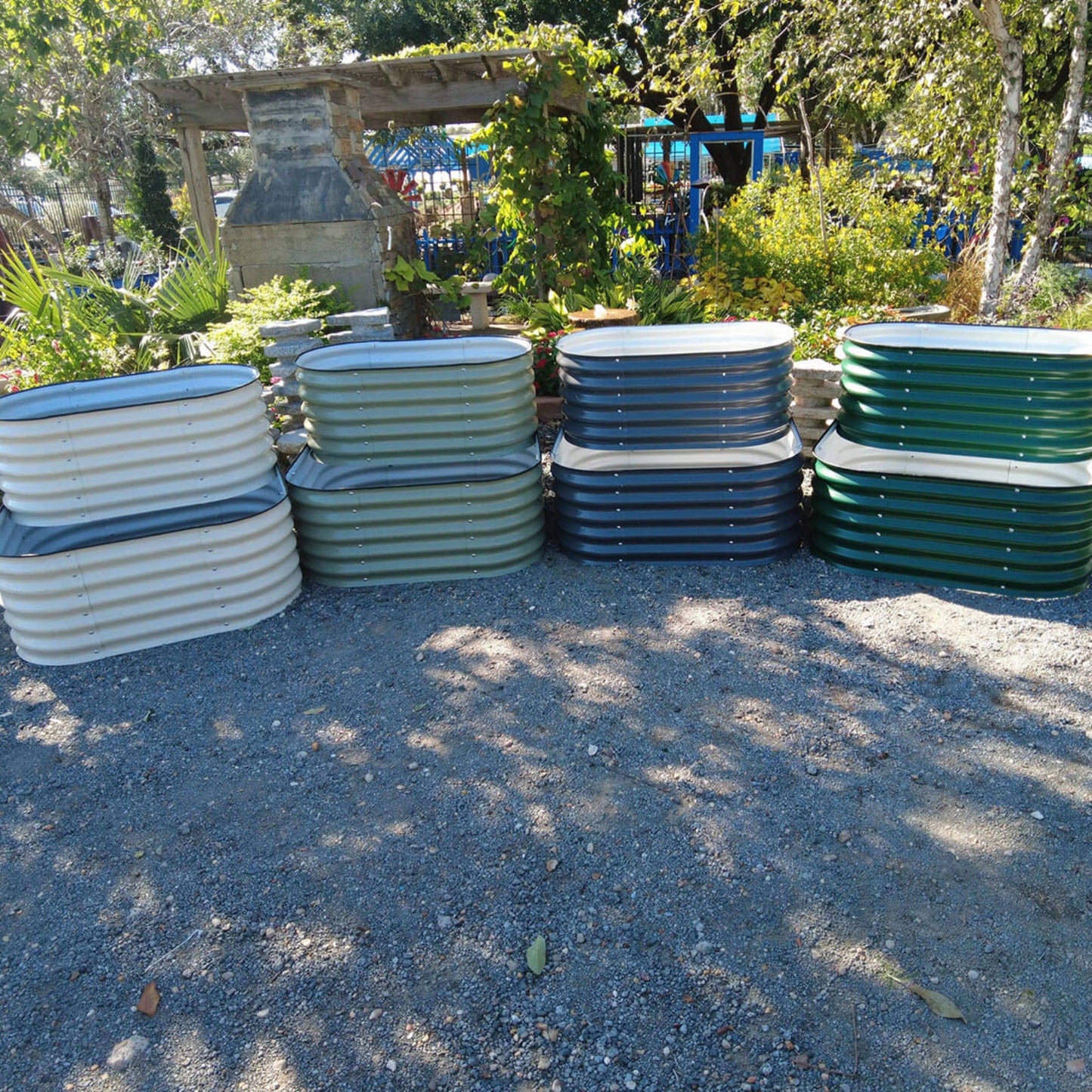 Four empty cascading metal raised garden beds, each showing the color options sitting on gravel in front of a garden landscape. 