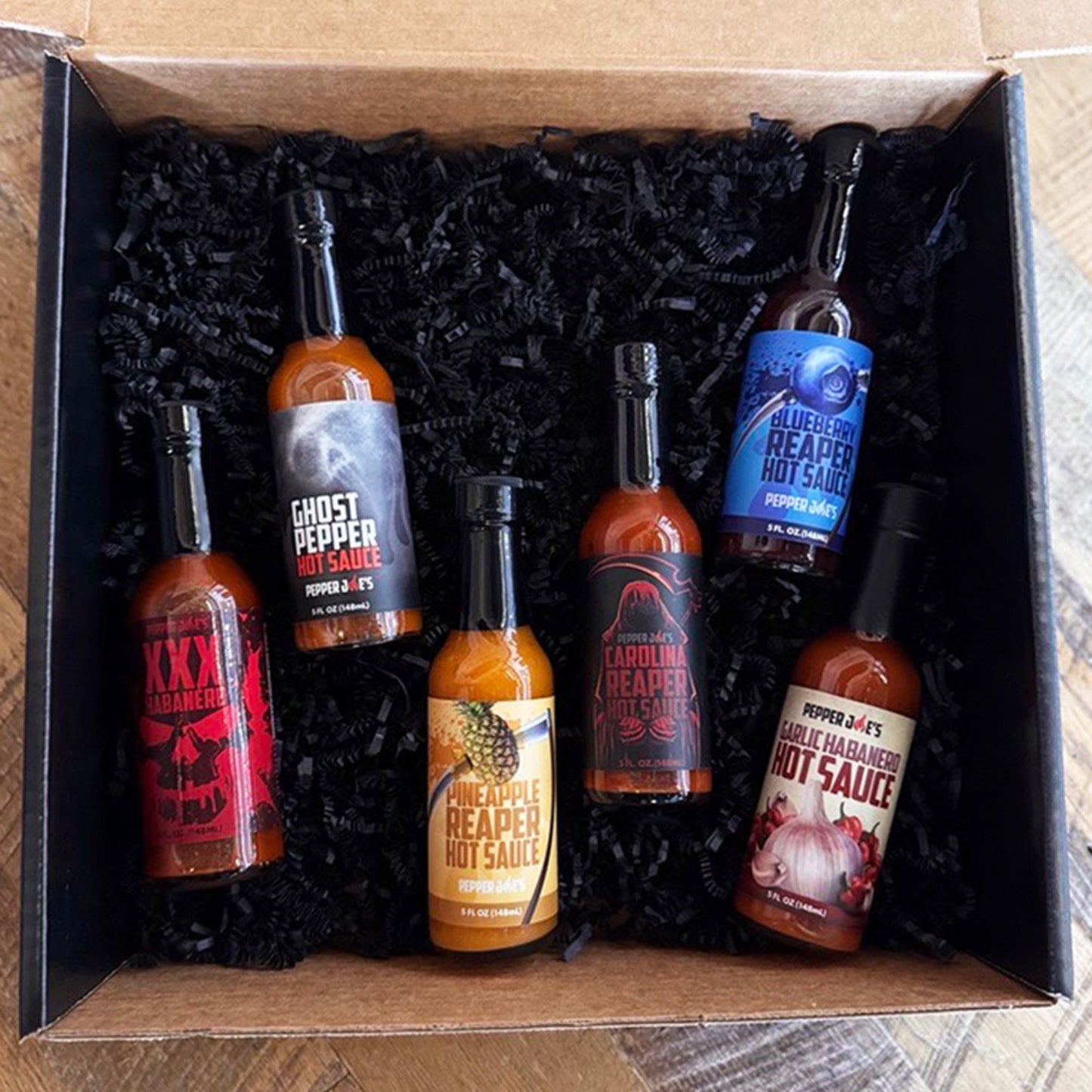 Pepper Joe's Hottesthot sauce gift box that includes one bottle each of XXX Habanero, Ghost Pepper, Pineapple Reaper, Carolina Reaper, Blueberry Reaper, and Garlic Habanero sauces inside gift box on a rustic background.