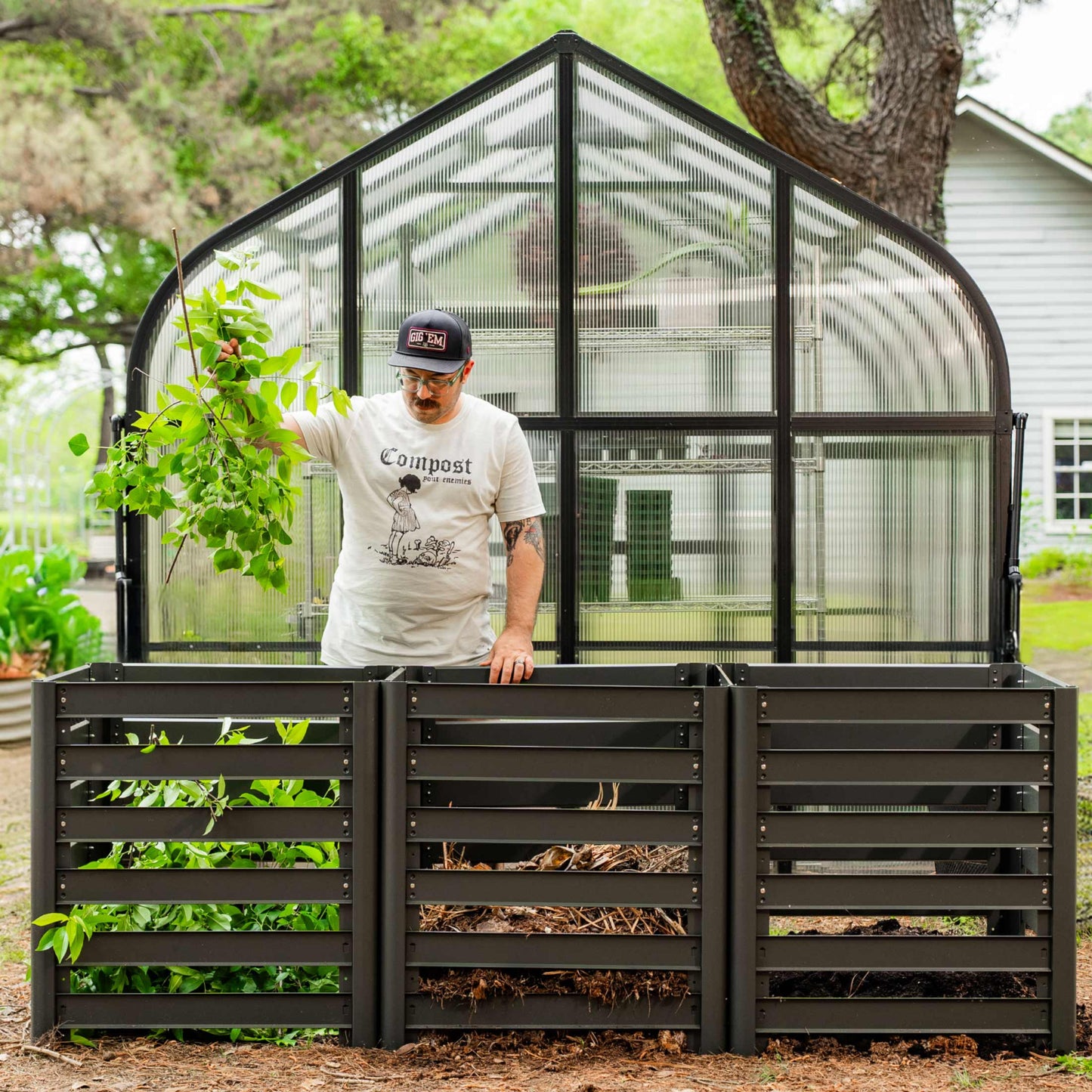 Person standing next to a modular compost bin with 3 bin configuration with a greenhouse in the background.