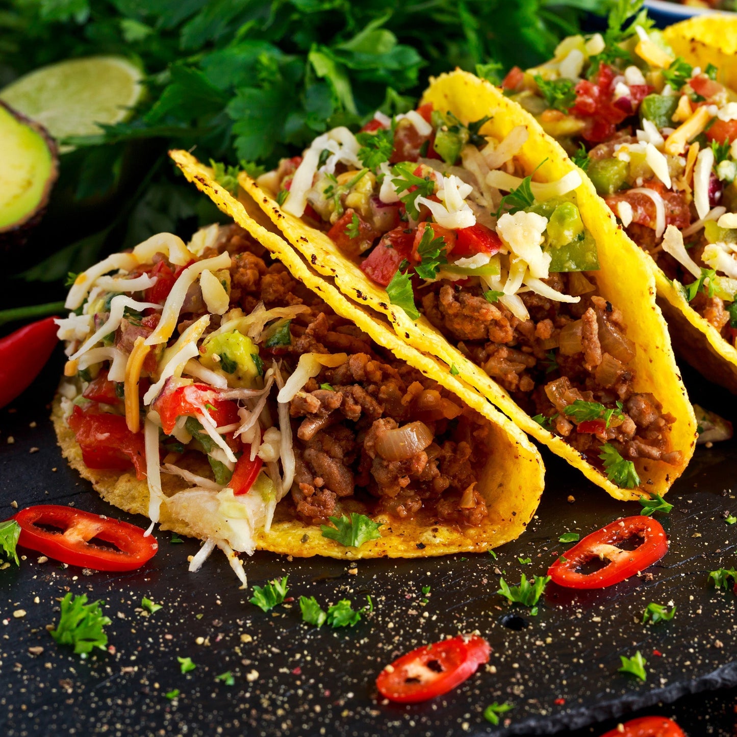 Two tacos with ground meat, cheese, and vegetables on a black surface with chili peppers and cilantro showing how to use the Scorpion Pepper Sea Salt in a meal.
