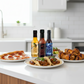 One bottle each of Pineapple Reaper, Carolina Reaper, and Blueberry Hot Sauce included in the Reaper Gift Box on a white kitchen counter beside a plate of pizza, plate of tacos, plate of fried chicken wings drizzled in the sauces.