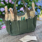 A green garden tool bag made of heavy waxed canvas, displayed on a wooden surface with gardening tools and paper gloves inside it, against a blurred floral background.
